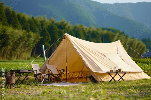 A beige canvas tent set up on the grass, with outdoor camping equipment such as folding chairs and tables next to it. In front of green mountains in summer, it has an ultra wide angle view