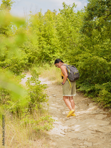Middle aged woman dressed in shorts walking along forest path with backpack. Attractive female traveler relaxing and enjoying nature on hiking trail. Concept of mental detox and physical health