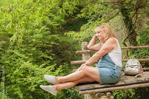 Mature woman sitting on wooden bench in summer forest. Attractive female traveler relaxing, meditation, breathing and enjoying nature on hiking trail. Concept of mental detox and physical health