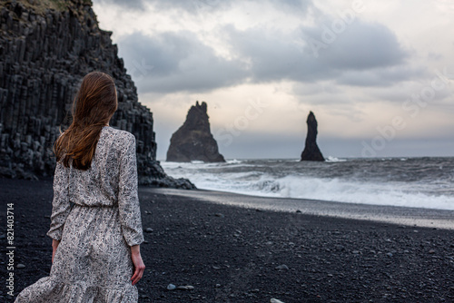 A beautiful brown-haired girl with beautiful flowing hair walks barefoot on a black beach in a dress near the ocean sea in Iceland in the summer photo