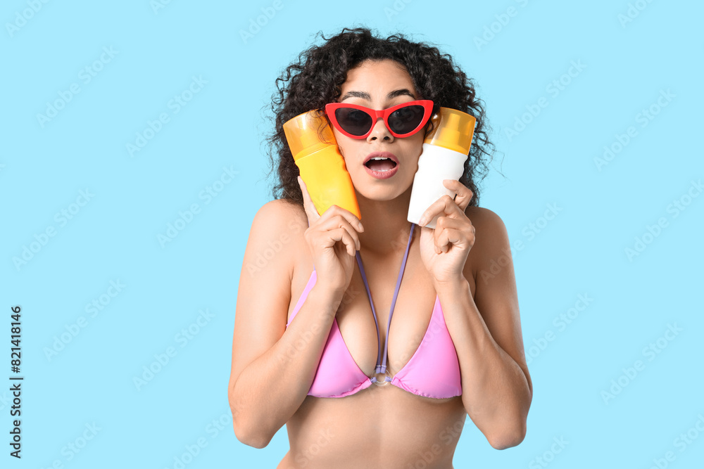Beautiful young African-American woman in swimsuit with bottles of sunscreen cream on blue background