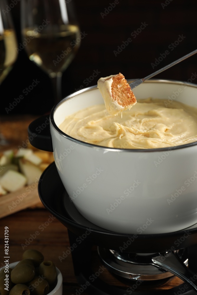 Dipping piece of bread into fondue pot with melted cheese at table, closeup