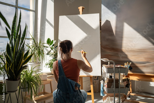 Creative artist woman starts to draw artwork on new white canvas on easel with brush at equipped modern art studio. Interested talented girl painter enjoy creative hobby for soul in painter workshop.