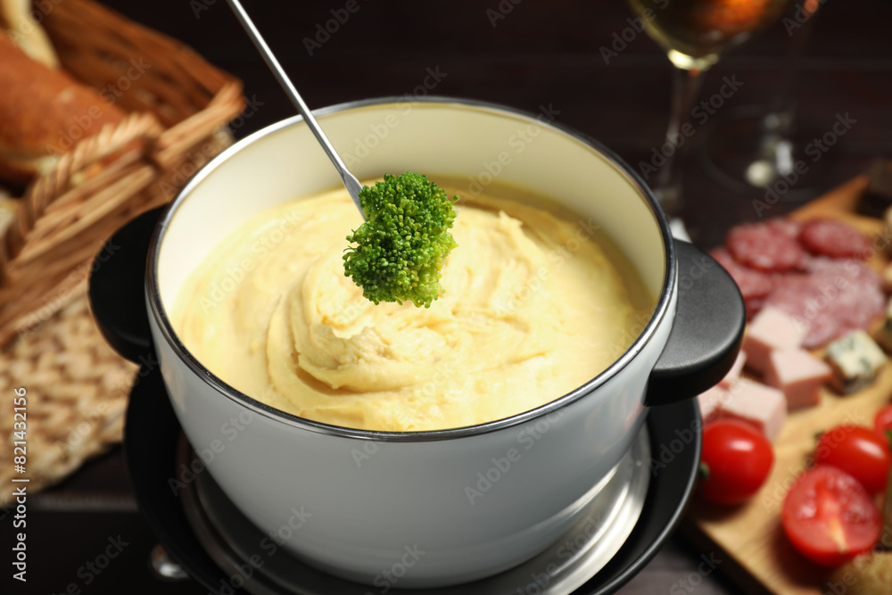 Dipping piece of broccoli into fondue pot with melted cheese at table with snacks, closeup Dipping piece of broccoli into fondue pot with melted cheese at table with snacks, closeup