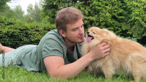 Young man is lying on green grass in park with his cute little Pomeranian Spits dog, guy play, hug and scratch his puppy at summer day