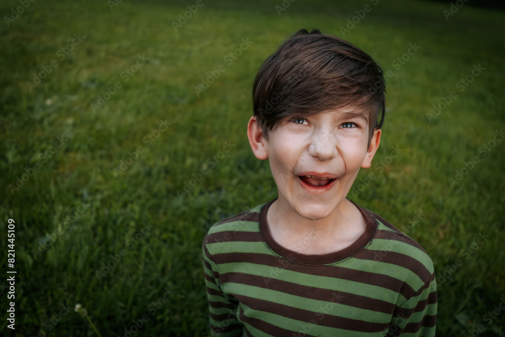 young boy with brown hair and blue eyes wearing a green and brown striped shirt makes a funny face while standing on a grassy field. The background is blurred young boy with brown hair and blue eyes wearing a green and brown striped shirt makes a funny face while standing on a grassy field. The background is blurred