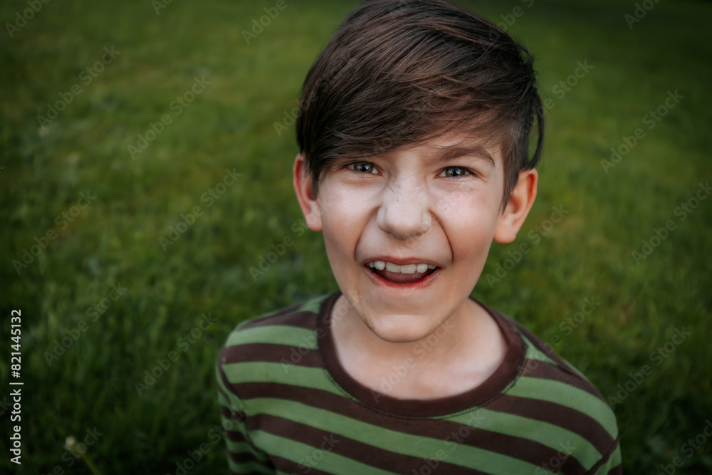 young boy with brown hair and blue eyes wearing a green and brown striped shirt makes a funny face while standing on a grassy field. The background is blurred young boy with brown hair and blue eyes wearing a green and brown striped shirt makes a funny face while standing on a grassy field. The background is blurred