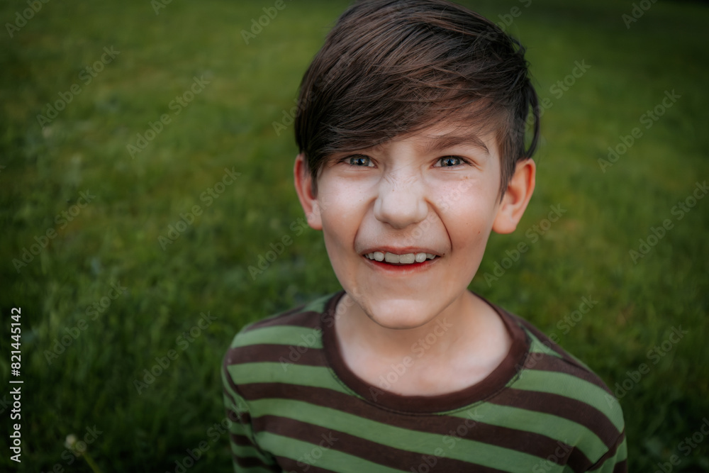 young boy with brown hair and blue eyes wearing a green and brown striped shirt makes a funny face while standing on a grassy field. The background is blurred young boy with brown hair and blue eyes wearing a green and brown striped shirt makes a funny face while standing on a grassy field. The background is blurred