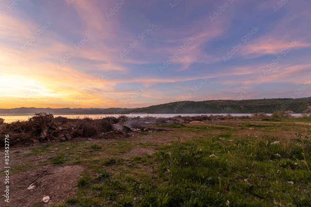  scene at the edge of a water body during sunset or sunrise, with a pile of burning logs and scattered garbage, highlighting the juxtaposition of natural beauty and environmental decay...