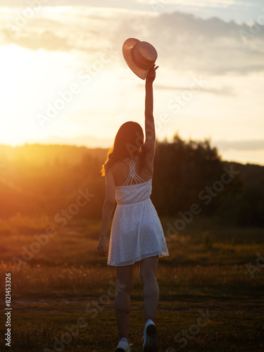 A happy girl in a white dress and a straw hat poses in a field at sunset. Summer time