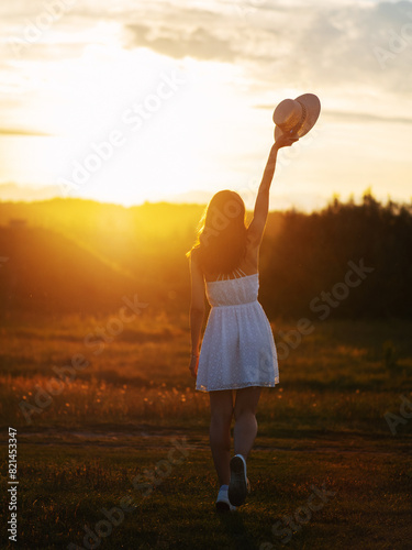 A happy girl in a white dress and a straw hat poses in a field at sunset. Summer time