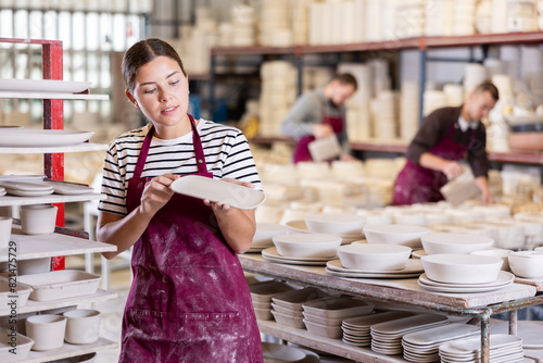 Interested professional young female ceramicist closely examining batch of slipcast pottery, inspecting technique and quality of workmanship in ceramic workshop.. photo