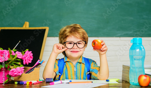 Healthy breakfast for pupils. Smiling cute child boy with apple during lunch break in classroom. Little schoolboy in glasses at desk in front of chalkboard eating apple. Healthy food menu for kids.