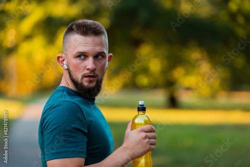 A young fit man listening to music on wireless headphones and drinking water or nutrition juice from a bottle after a run or workout in a summer park active healthy lifestyle.