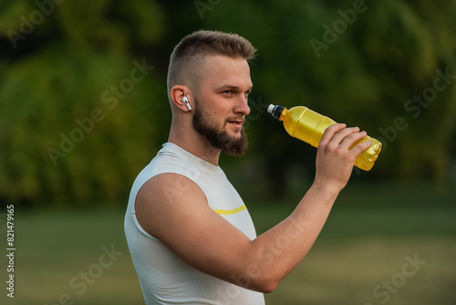 A sporty young man listening to music on wireless headphones and drinking water or nutrition juice from a bottle after a run or workout in a summer park active healthy lifestyle..