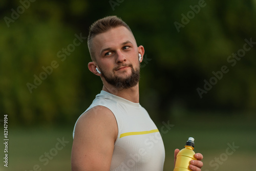 A young happy fit man listening to music on wireless headphones and drinking water or nutrition juice from a bottle after a run or workout in a summer park active healthy lifestyle.