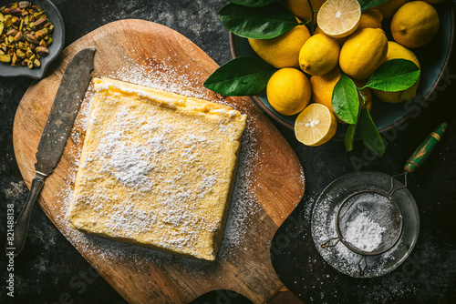 Lemon cake, unsliced, on wood cutting board with cake knife, powdered sugar, and bowl of fresh lemons photo