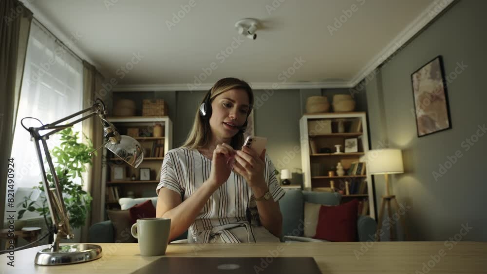 Happy attractive woman with headset using mobile phone while sitting at desk with a laptop in front of her