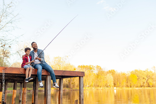 African American man and child sitting on a wooden pier holding fishing rods on the river, dad teaching his son to fish in the lake, family resting and relaxing on the weekend