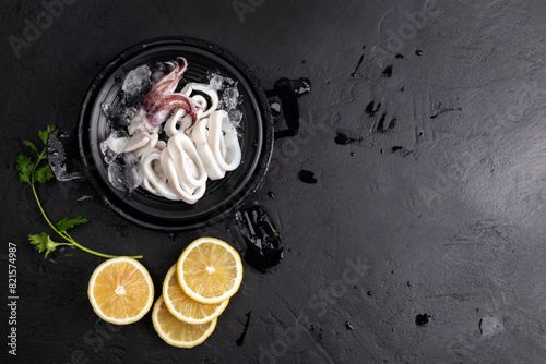 Fresh raw squid. Sliced ​​raw squid placed on a black plate with ice, lemon slices. Top view and copy space for your text.
