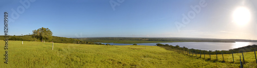 Panoramic photo of a lush green hay plantation under rural blue sky, grass texture and beautiful lake in the background. Beautiful morning light on green grass farm. Pedra do Cavalo Dam Lagoon