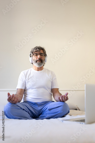 Vertical portrait of mature man sitting on bed at home following online meditation classes using laptop and headphones.