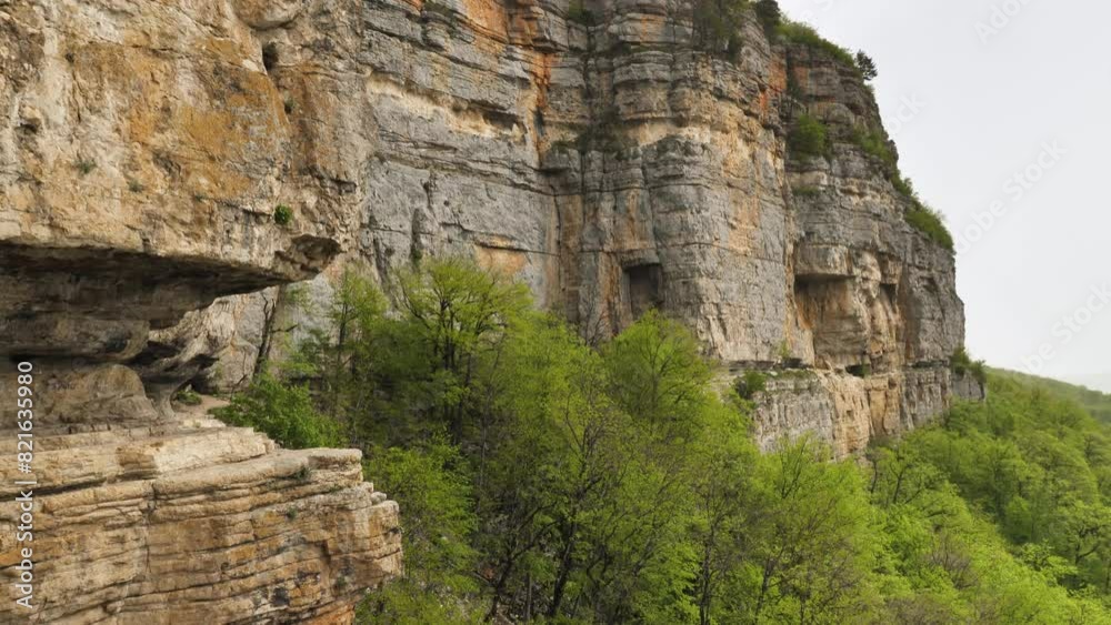 A towering cliff with layers of sedimentary rock in beige and light brown colours. Trees and green foliage are visible at base and slope of the cliff. The cliff is part of a large mountainous area A towering cliff with layers of sedimentary rock in beige and light brown colours. Trees and green foliage are visible at base and slope of the cliff. The cliff is part of a large mountainous area