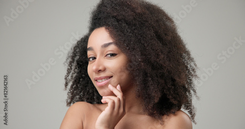 Beauty and healthcare concept - beautiful African American woman with curly afro hairstyle and clean, healthy skin touches her cheek and face with her hand, posing and looking at the camera
