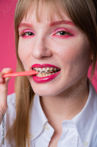 Portrait of a young woman with braces and bright makeup chewing gum on a pink background. Vertical photo.
