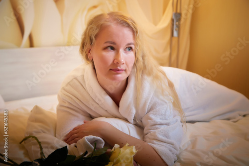 A blonde girl in a white terry bathrobe in a hotel room with flowers. Wedding travel, Weekend and vacation, body care
