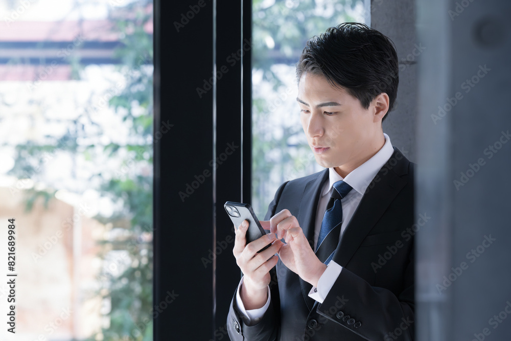 窓際で真剣な表情でスマホを見る(検索や操作)、タップするスーツを着たビジネスマン 窓際で真剣な表情でスマホを見る(検索や操作)、タップするスーツを着たビジネスマン