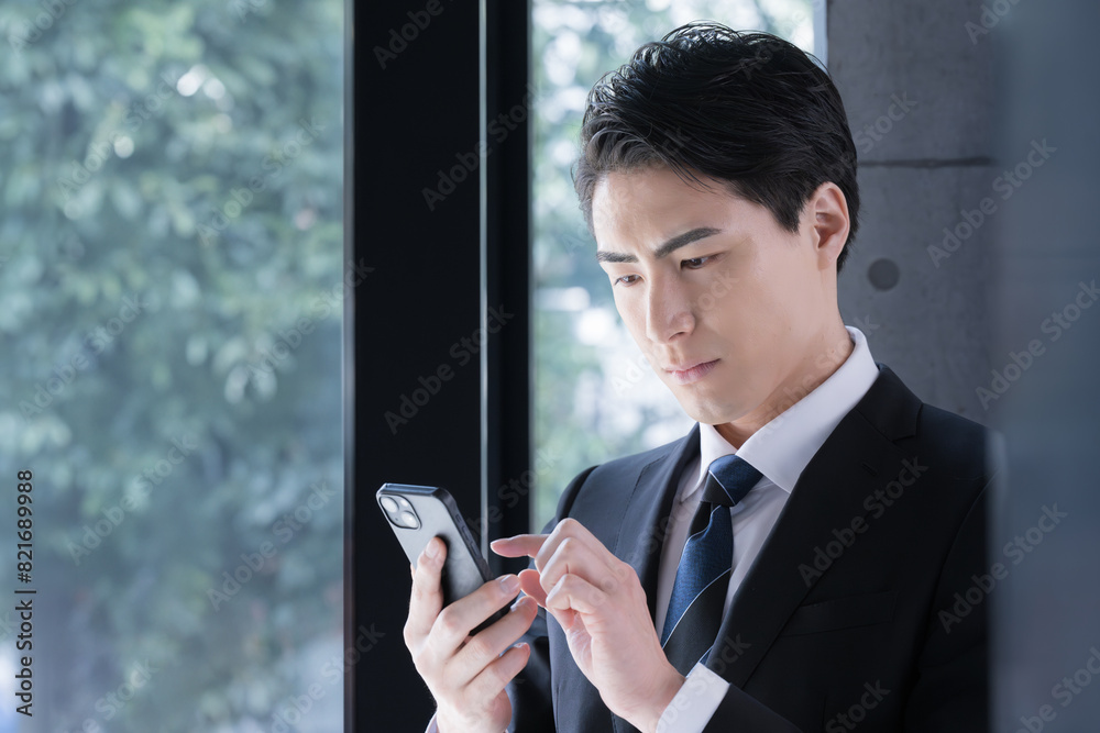 窓際で真剣な表情でスマホを見る(検索や操作)、タップするスーツを着たビジネスマン 転職などのイメージにも 窓際で真剣な表情でスマホを見る(検索や操作)、タップするスーツを着たビジネスマン 転職などのイメージにも