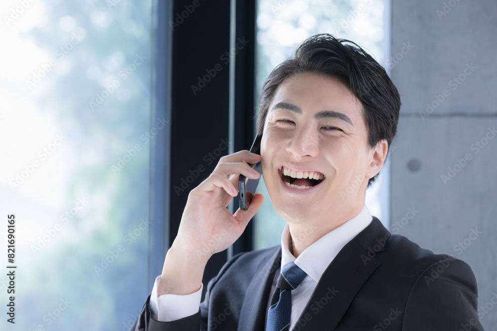 窓際で笑顔で電話(通話)をするスーツを着た男性ビジネスマン　カメラ目線　逆光の中大笑いする成功のイメージ
