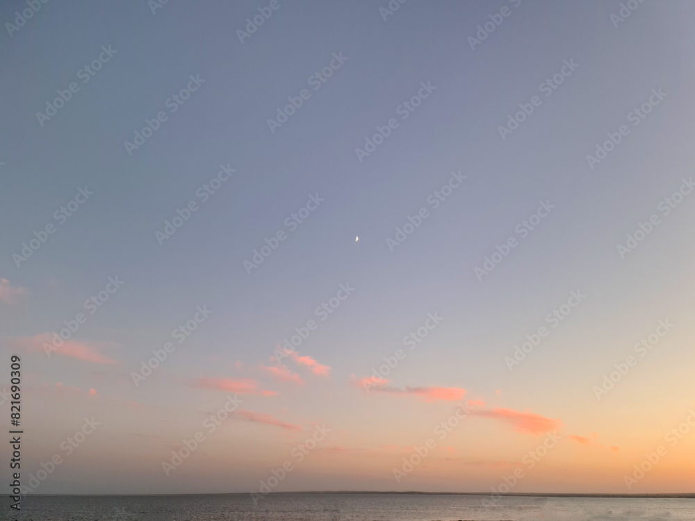 Little moon hangs in the sky over the ocean during dusk, accompanied by a vibrant afterglow. A beautiful natural landscape at sunset.