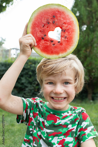 cheerful boy eats a watermelon, cutting out a heart in a round piece of watermelon and putting it on his head. Hello summer, good mood, delicious vitamin food. The joy of childhood