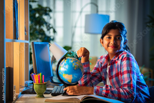 Asian Indian girl child studying at home on study table with computer, books, Globe model, victory trophy