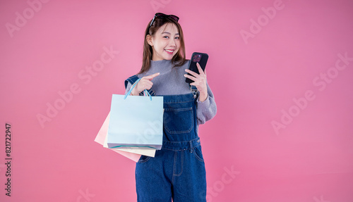 Portrait happy beautiful shopaholic asian woman carry shopping bags, winter fashion sale model Asia girl with mobile phone, outlet department store advertise concept isolated pink banner background