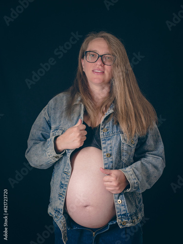 A mom-to-be in late pregnancy wearing an unbuttoned denim jacket. Belt portrait of a pregnant woman on a dark background. Joy and anticipation of motherhood.