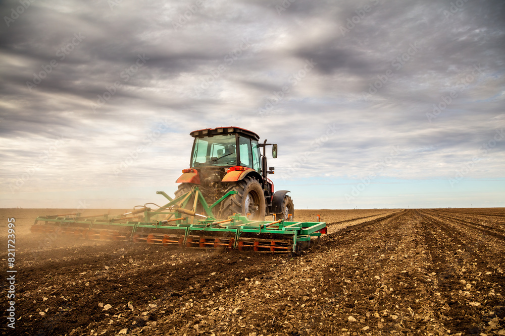 Fototapeta premium Powerful tractor tills the soil, preparing the land for planting under a dramatic sky