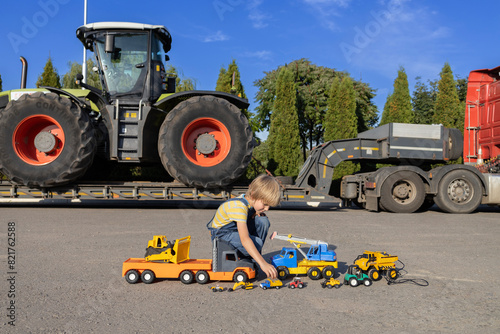 7-year-old boy plays with toy cars while a huge tractor on a transport trailer stands behind him. interesting childhood of a child, games in the profession
