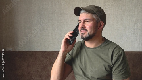 A man with a beard is calling on the phone. In hand there is a black smartphone. Green baseball cap and T-shirt. Military. In room. Sitting on a brown sofa against the background of a wall.