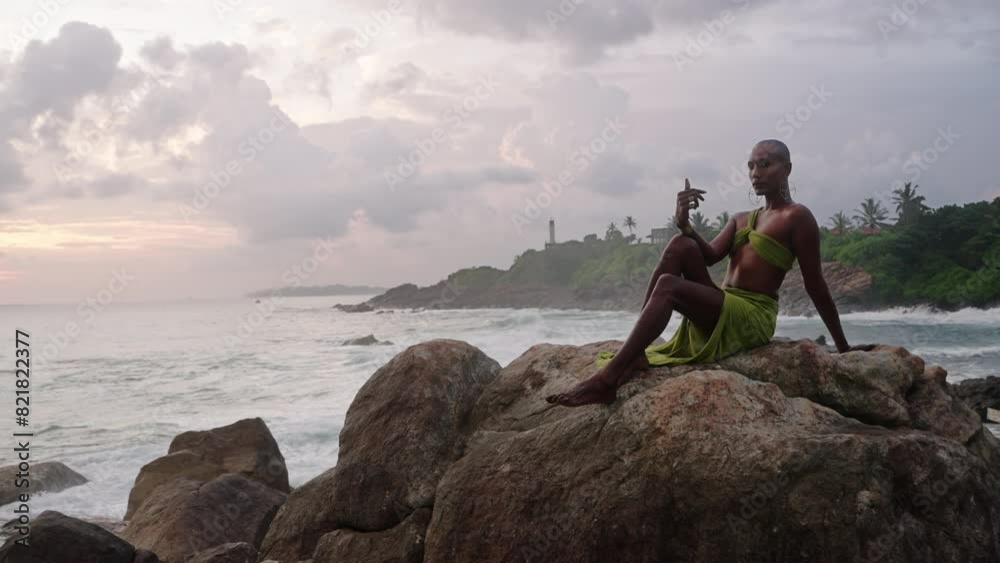 LGBTQIA black person in open dress, jewelry sits on rock on stunning ocean coastline sunset. Gender fluid ethnic divine model in seductive outfit fluttering in the wind against dramatic sky. LGBTQIA black person in open dress, jewelry sits on rock on stunning ocean coastline sunset. Gender fluid ethnic divine model in seductive outfit fluttering in the wind against dramatic sky.