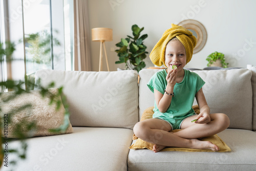 Smiling girl sitting on sofa and eating cucumber at home photo