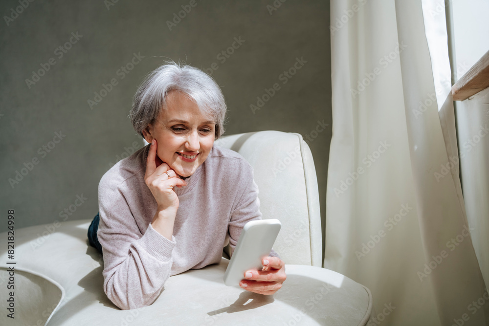 Smiling mature woman video calling through smart phone on couch at home Smiling mature woman video calling through smart phone on couch at home
