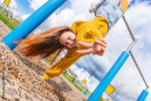 Cheerful girl hanging upside down on gymnastics bar and showing peace sign