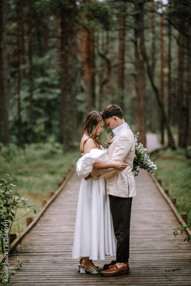 Riga, Latvia, - August 26, 2024 - Bride and groom embracing on a wooden path in a lush forest, with the bride holding a bouquet...