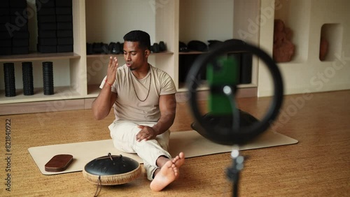 Black male blogger sitting on yoga mat with Sadhu Board and glucophone, talking about practice in front of mobile phone with ring light while filming class for fitness channel. Shooting in slow motion