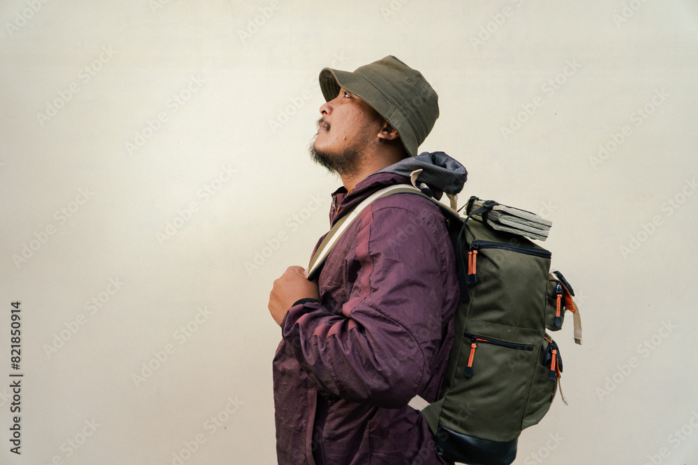 Half body portrait of adult Asian man with beard and mustache posing like an adventurer. Model in hiking clothes with jacket, bucket hat, carrier bag isolated on beige background. Half body portrait of adult Asian man with beard and mustache posing like an adventurer. Model in hiking clothes with jacket, bucket hat, carrier bag isolated on beige background.
