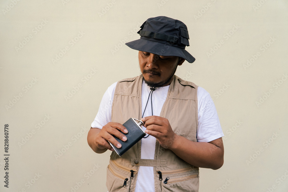 Macho asian man with beard and mustache wearing safari clothes isolated on beige background. Half body portrait of adult Southeast Asian man posing wearing vest, bucket hat and hip flask