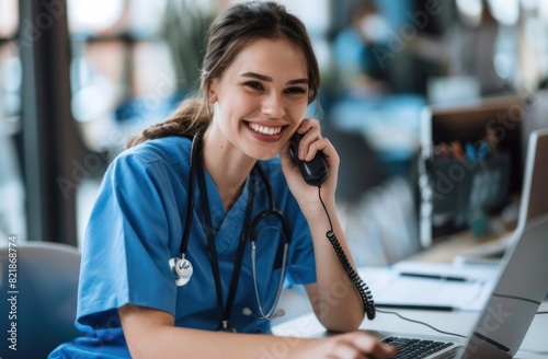 Young female nurse in blue scrubs talking on the phone while sitting at her desk with laptop.